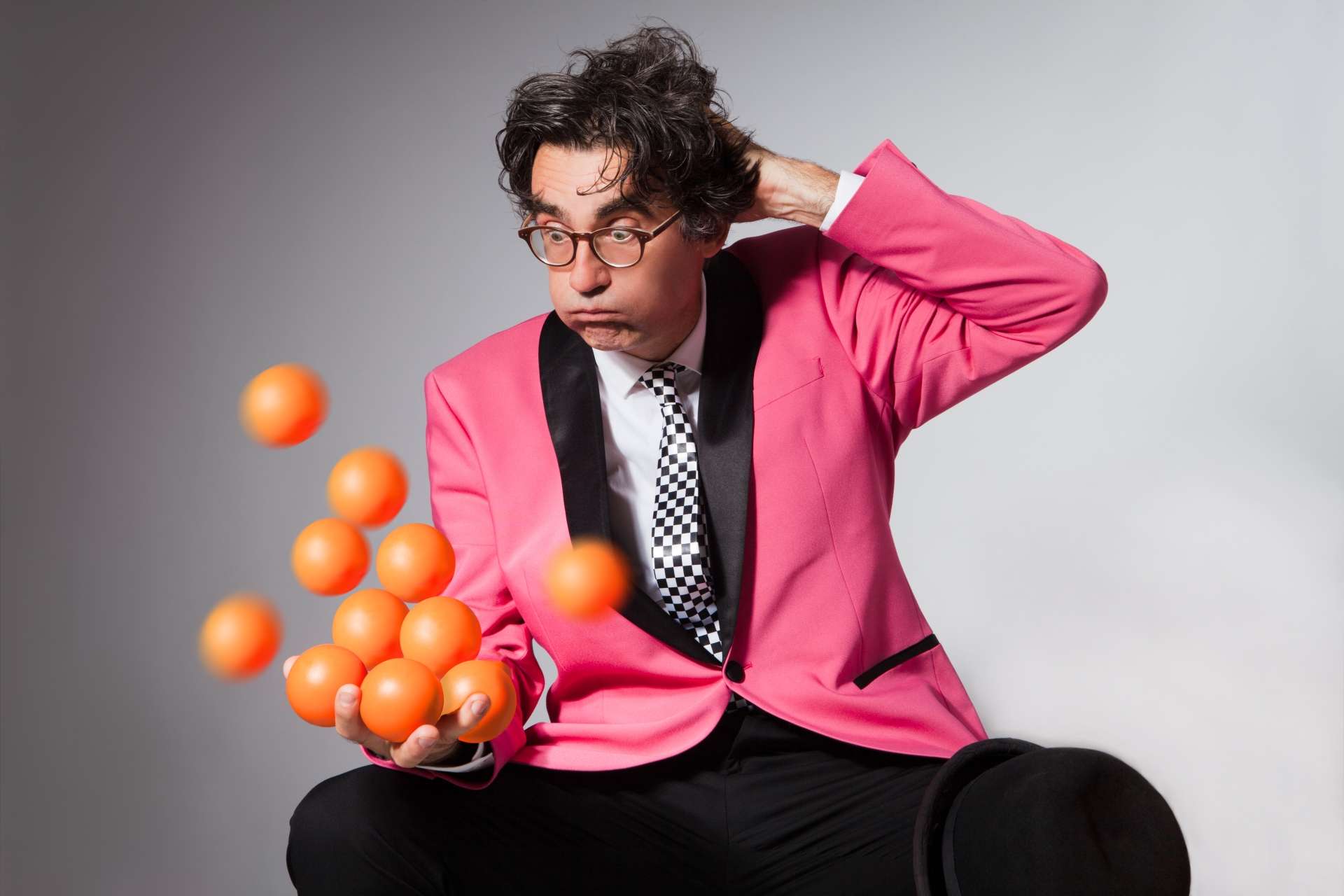 Un homme vêtu d'un costume rose lance un dizaine de balle orange dans les airs avec une tête embarrassé en se grattant la tête.