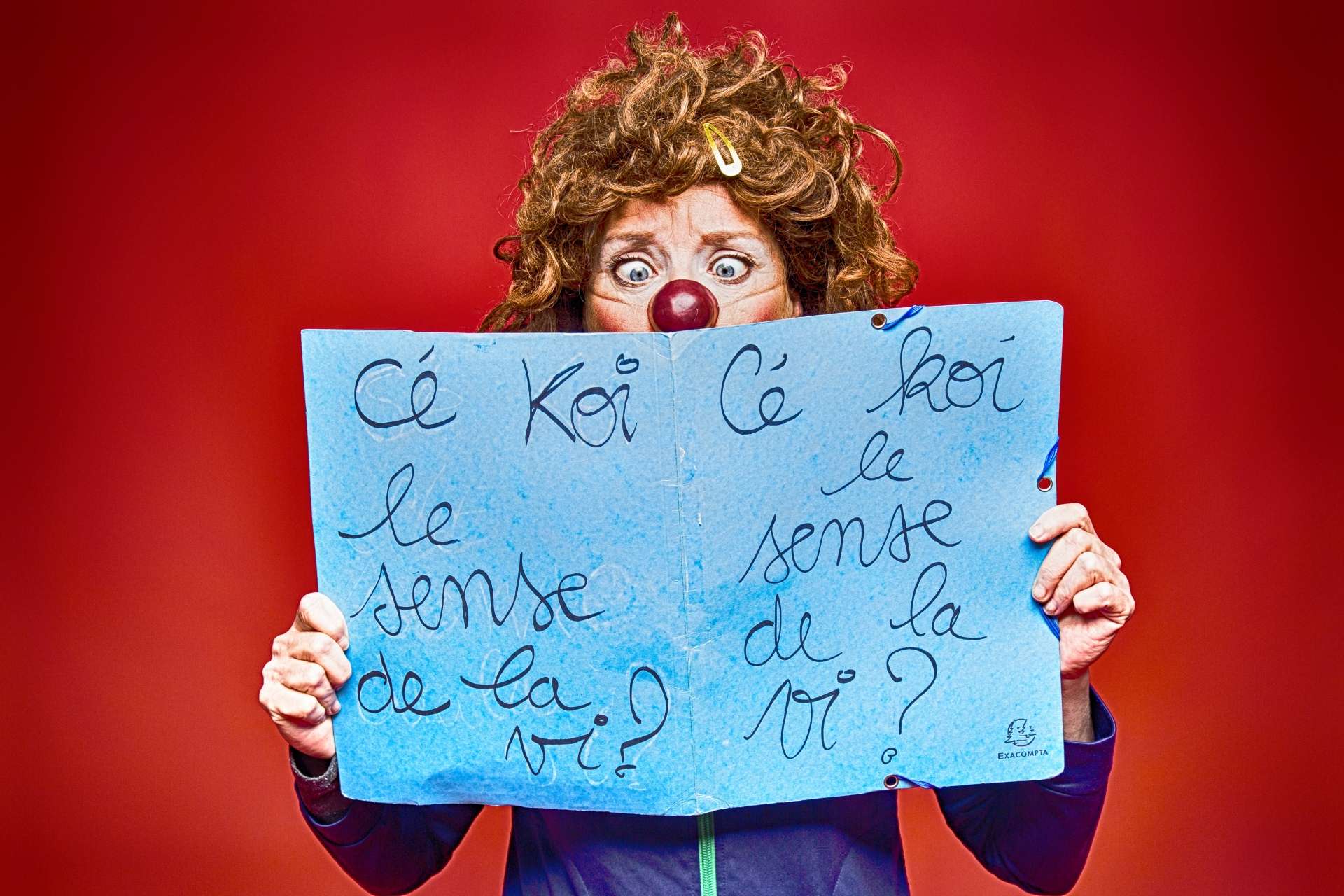 Une femme déguisée en clown sur un fond rouge tient une affiche avec écrit "Cé koi le sense de la vi ?"
