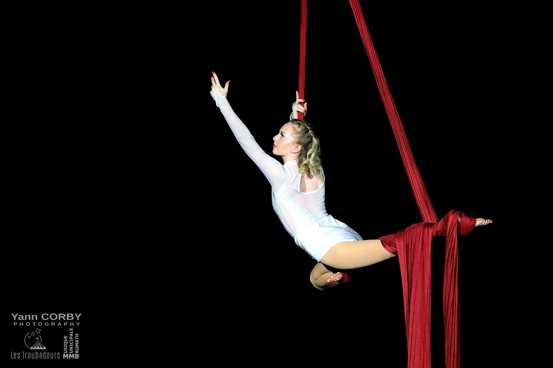 Une femme habillée en blanc fait des acrobaties dans les airs sur un tissus rouge.
